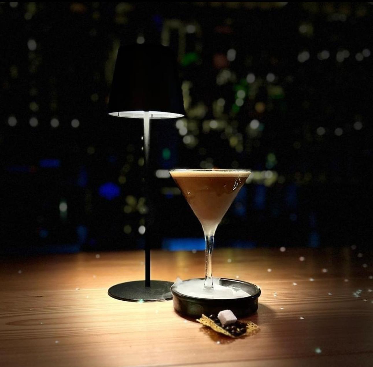 Martini glass with a cocktail beside a lit table lamp on a wooden surface in dim lighting.