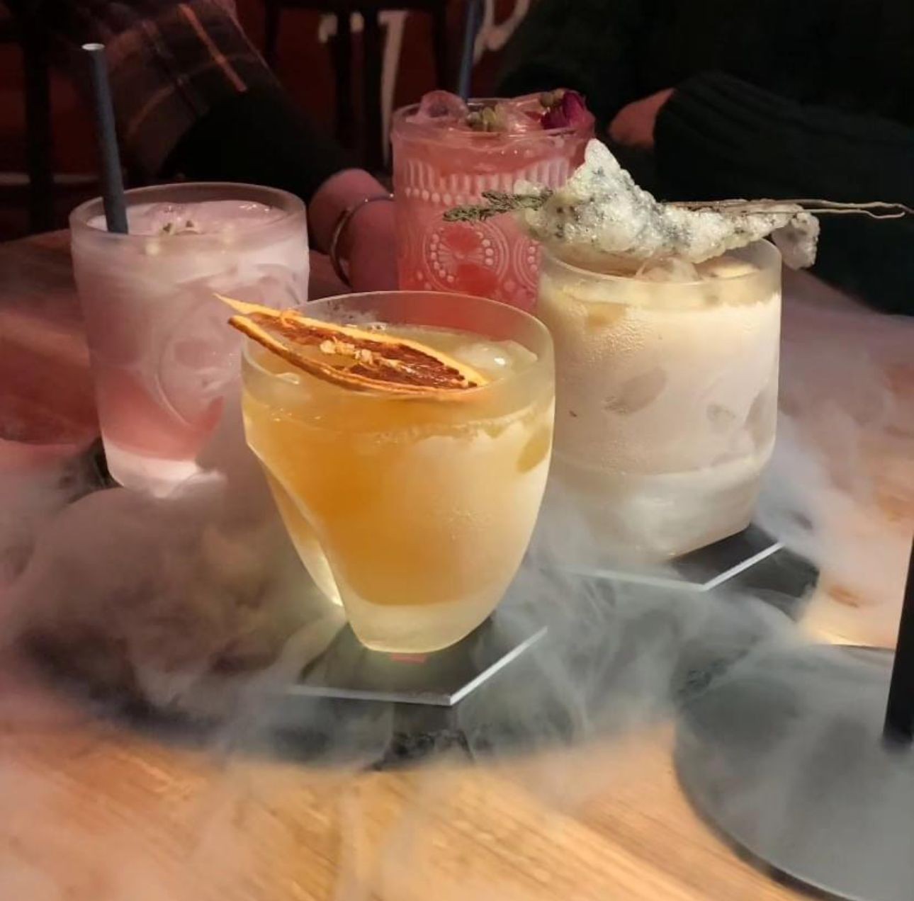 Assorted colorful cocktails with dry ice fog on a wooden table.